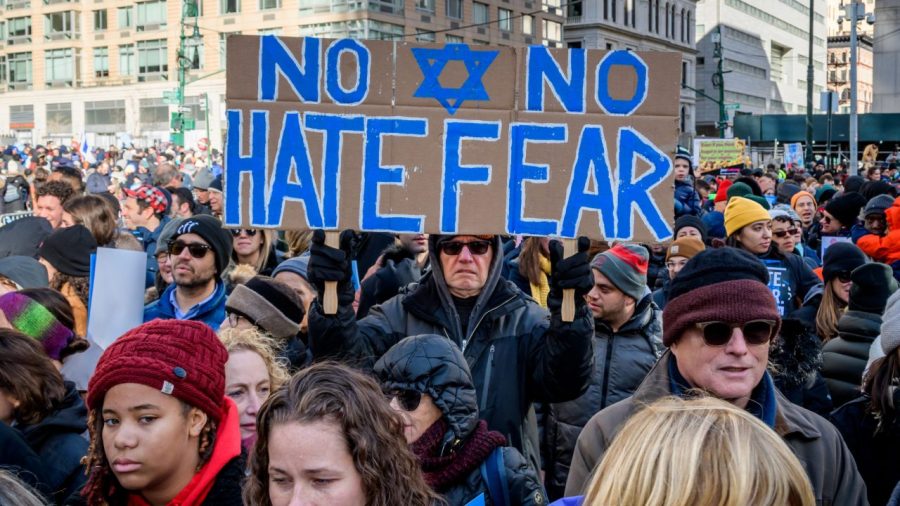 A group of people in front of a city backdrop, with a cardboard sign featuring a Star of David; the sign reads, NO HATE NO FEAR.